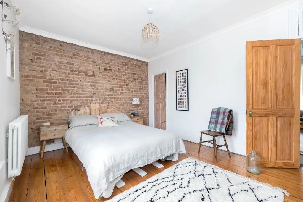 Bedroom before renovation with warm orange wood flooring, white walls, and original exposed brick wall