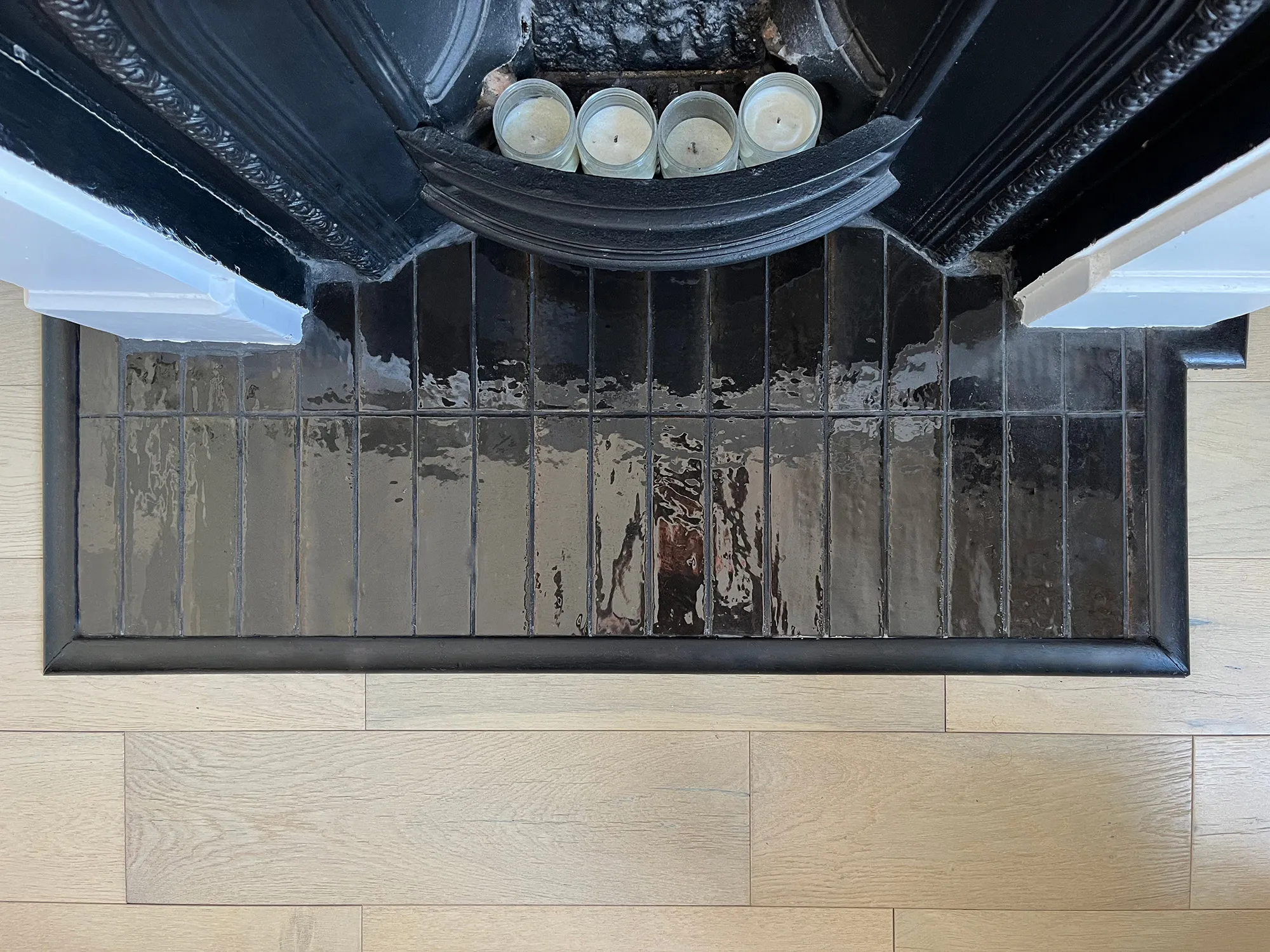 Top down view of black glossy zellige hearth tile next to light wood flooring and cast iron fireplace