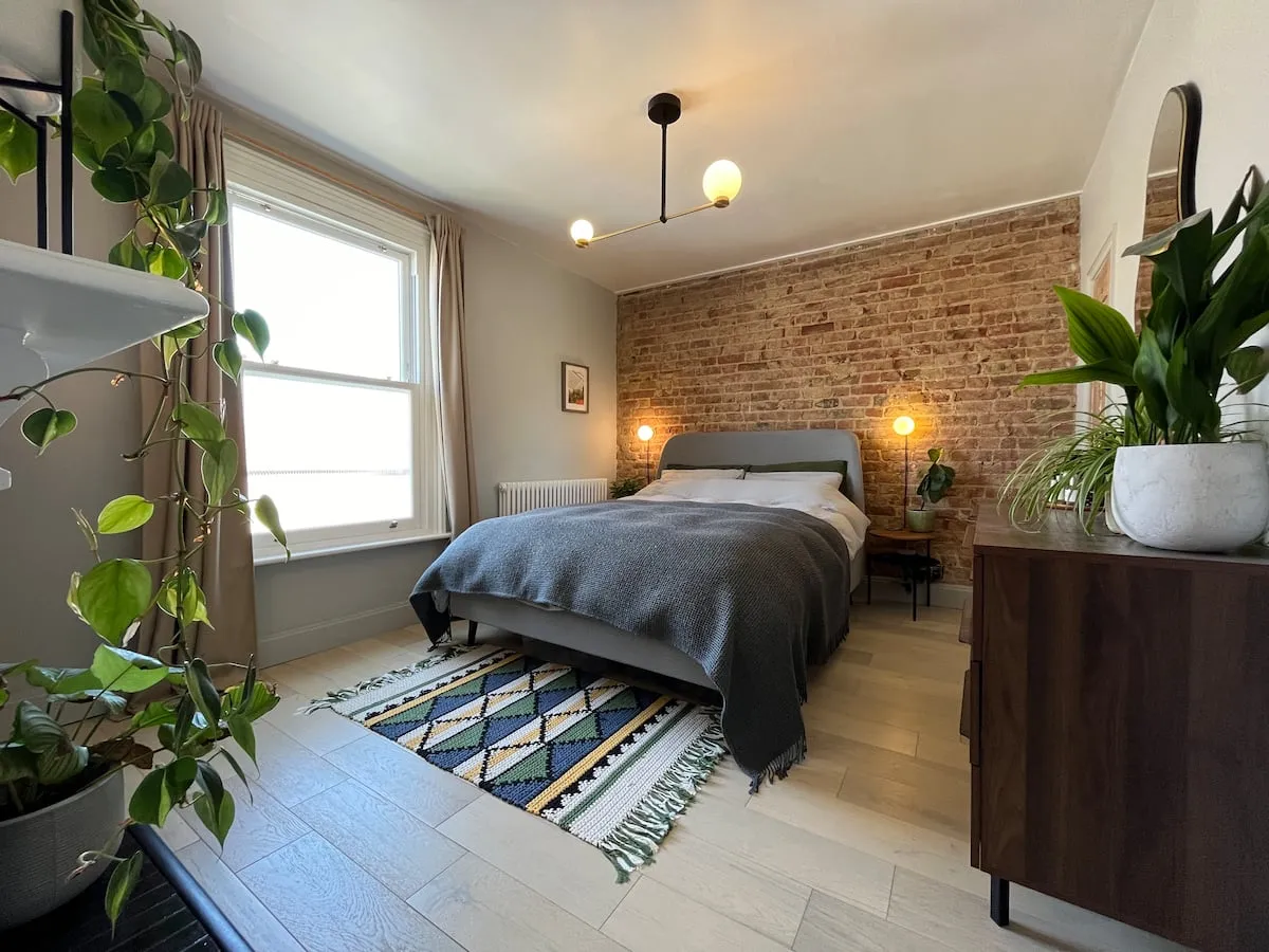 Renovated modern bedroom featuring exposed brick wall, light oak floors, dark grey bed, and potted houseplants